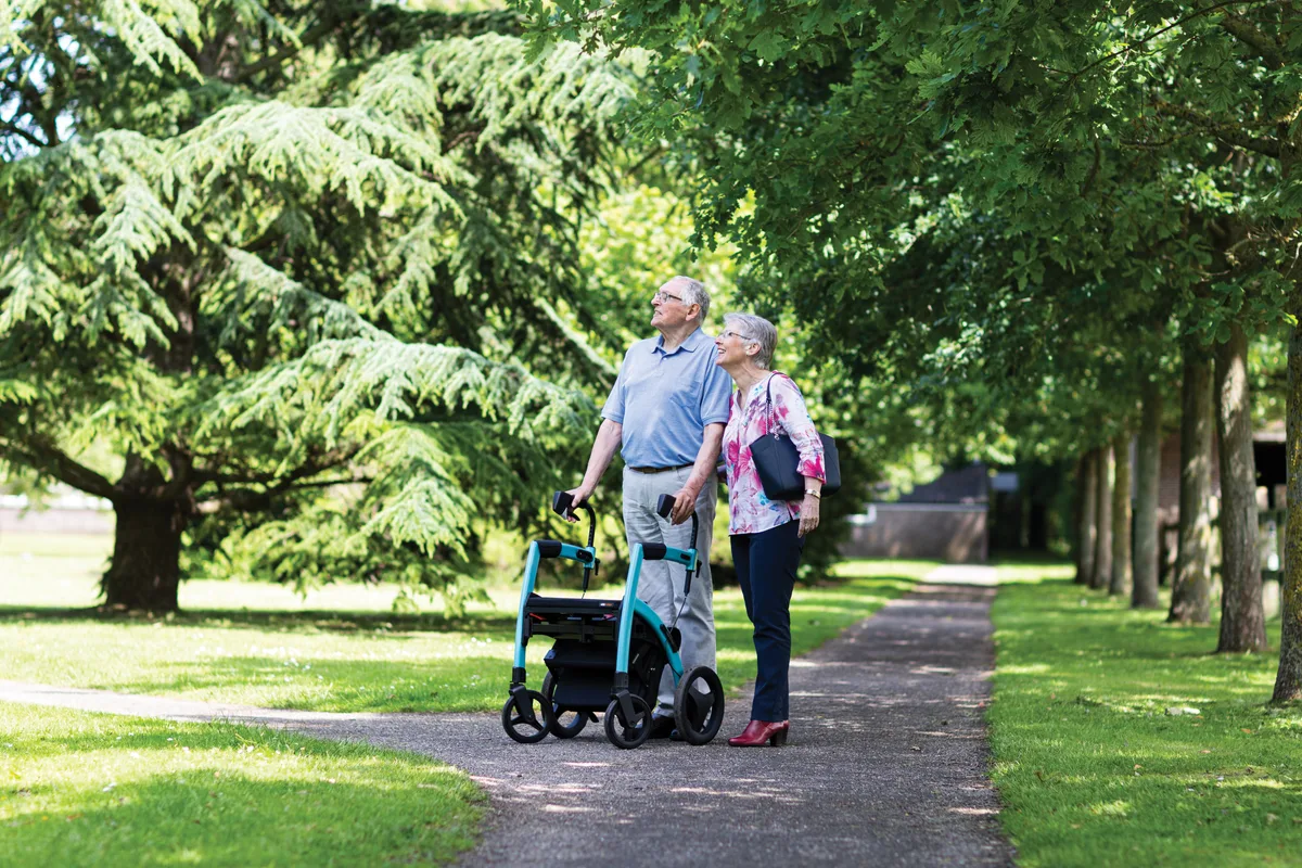 Two older adults walking together on a tree-lined path, one using a walker